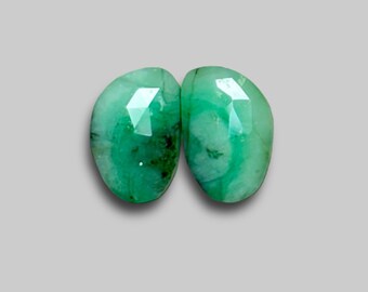 May include: Two faceted emerald gemstones with a vibrant green color. The gemstones are oval-shaped and have a polished, reflective surface. The background is a solid, neutral gray.