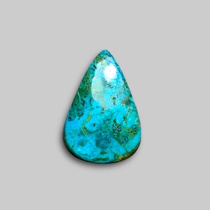 May include: A teardrop-shaped gemstone with a polished surface. The stone features a blend of turquoise, teal, and hints of green and brown. The gemstone's unique pattern and color variations are visible. The stone is set against a neutral gray background.