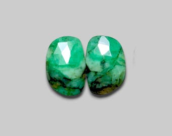 May include: Two faceted emerald gemstones with a vibrant green color and visible inclusions. The gemstones have an oval shape and are set against a neutral gray background. The facets reflect light, creating a sparkling effect.