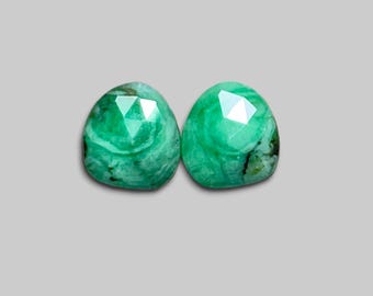 May include: Two faceted, oval-shaped emerald gemstones. The gemstones are a vibrant green color with internal swirls and dark inclusions. The facets catch the light, creating a sparkling effect. The stones are set against a neutral gray background.