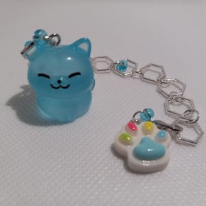 Pode incluir: Um amuleto de gato azul claro com um rosto sorridente, preso a uma corrente prateada com um amuleto de pata azul claro com detalhes coloridos.