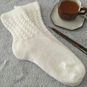 Peut inclure: Une paire de chaussettes blanches en tricot avec un motif de câble. Les chaussettes sont posées sur une surface grise et duveteuse.