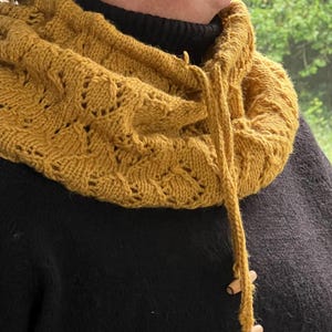 May include: Mustard yellow knitted cowl with a drawstring and wooden toggles. The cowl features a textured, openwork design. The drawstring is made of the same yarn as the cowl. The cowl is worn over a black turtleneck sweater.