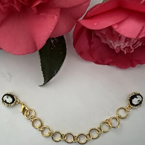 Op de afbeelding: Een goudkleurige kettingarmband met twee ovale cameohangers. De cameeën tonen een wit profiel tegen een zwarte achtergrond. De armband wordt getoond met roze bloemen.