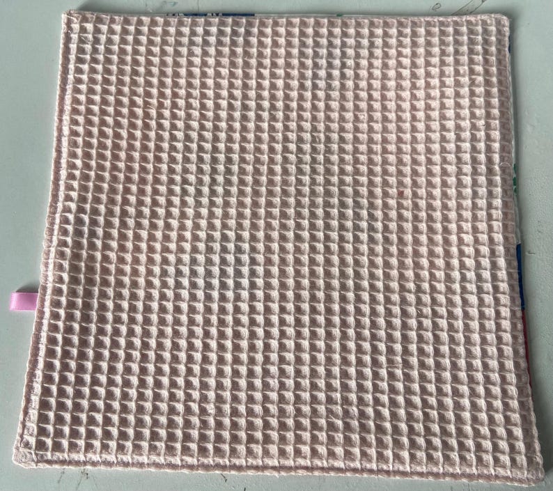 May include: A pale pink waffle weave kitchen towel. The towel is made of a soft, absorbent material and is perfect for drying dishes, hands, and surfaces.