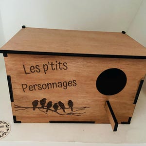 Puede incluir: Una casita de pájaros de madera con un borde negro y una abertura redonda. La casita está decorada con el texto "Les p'tits Personnages" y una silueta de pájaros en una rama.