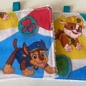 Può includere: Due elementi quadrati in tessuto con bordo bianco e passante turchese. Ognuno presenta un personaggio diverso di Paw Patrol su uno sfondo geometrico colorato. Uno ha Chase, l'altro Rubble. Lo sfondo ha forme blu, gialle e rosse.