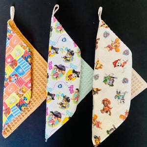 Puede incluir: Tres baberos triangulares para bebés con respaldo de tejido de gofre. Un babero presenta un patrón colorido de personajes de dibujos animados, mientras que los otros muestran un patrón repetido de cachorros de dibujos animados. Cada babero tiene un lazo para colgar.