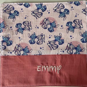 May include: A pink and white fabric pouch with a repeating pattern of blue cartoon characters. The pouch has a red flap with the name "Emmy" embroidered on it.
