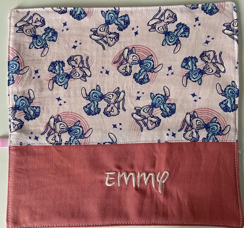 May include: A pink and white fabric pouch with a repeating pattern of blue cartoon characters. The pouch has a red flap with the name "Emmy" embroidered on it.