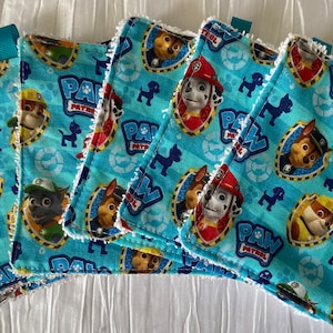 Può includere: Un set di cinque elementi quadrati in tessuto azzurro con i personaggi di Paw Patrol. Ogni elemento ha un bordo bianco e un piccolo anello turchese. Il tessuto presenta un motivo ripetuto del logo e delle immagini dei personaggi di Paw Patrol.