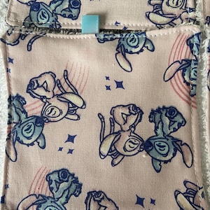 Può includere: Tessuto rosa con un motivo ripetuto di personaggi dei cartoni animati blu e bianchi. I personaggi sono una versione stilizzata di Stitch del film Disney Lilo & Stitch. Il tessuto è cucito a forma di quadrato con un nastro blu chiaro cucito sulla parte superiore.