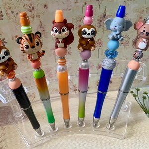 Peut inclure: Six stylos présentés sur un support transparent, chacun surmonté d'une perle en silicone représentant un personnage. Les personnages incluent un paresseux, un tigre, un écureuil, un autre paresseux, un éléphant et une souris, avec des perles colorées.