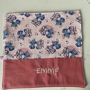 May include: Pink fabric pencil case with a blue and white cartoon pattern of Stitch from Lilo & Stitch. The case has a pink flap with the name "EMMY" embroidered on it.