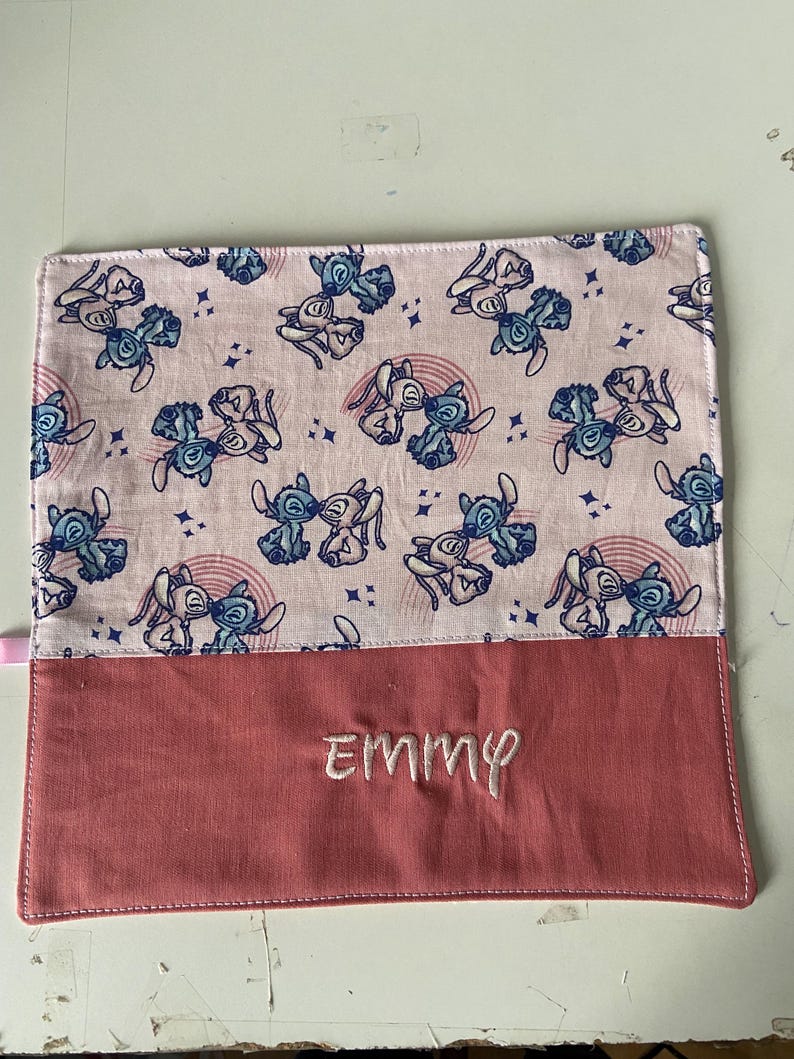 May include: Pink fabric pencil case with a blue and white cartoon pattern of Stitch from Lilo & Stitch. The case has a pink flap with the name "EMMY" embroidered on it.