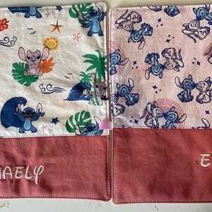 May include: Two pink and white fabric squares with a blue cartoon character pattern. The squares have a red fabric bottom with the names "Maely" and "Emmy" embroidered on them.