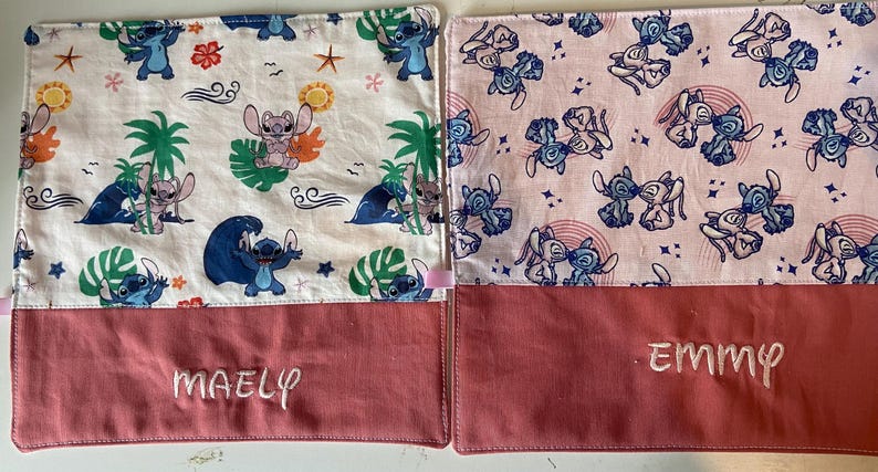May include: Two pink and white fabric squares with a blue cartoon character pattern. The squares have a red fabric bottom with the names "Maely" and "Emmy" embroidered on them.