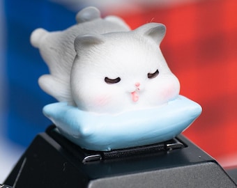 May include: A cute, gray and white cat figurine resting on a light blue pillow. The cat has closed eyes and pink cheeks. The figurine is on top of a black camera. A red, white, and blue object is in the background.