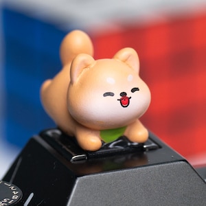 May include: A small, tan Shiba Inu figurine with a green collar and a happy expression sits atop a black camera. The background is blurred, with hints of red, white, and blue.