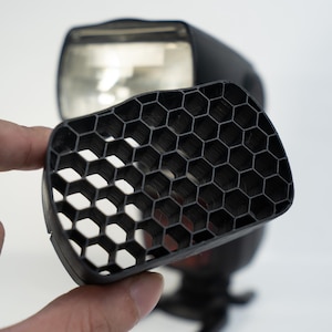 May include: A black honeycomb grid light modifier is held in front of a black camera flash. The grid is made of a series of hexagonal cells. The flash is black with a silver reflector.