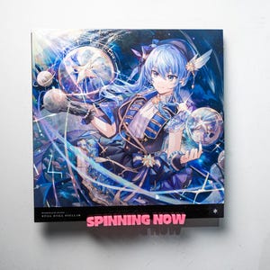May include: A vinyl record album cover featuring an anime-style illustration of a character with blue hair and a blue and gold outfit. The artwork has a celestial theme with stars and planets. The text "SPINNING NOW" is displayed in pink.