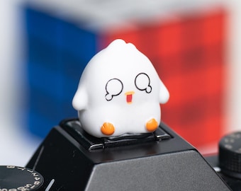 May include: A small, white, cartoon-style chick figurine with black eyes, orange feet, and a red beak. The chick is perched on a black camera. A blurred red, white, and blue background is visible.