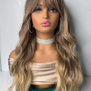 May include: A long, wavy wig with a dark brown to blonde ombre effect. The wig features a fringe and is styled on a mannequin head. The mannequin is wearing a gold-colored top, a rhinestone choker, and large hoop earrings.