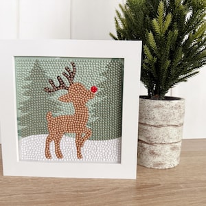 May include: A framed Christmas art piece featuring a reindeer design. The reindeer is brown with a red nose and brown antlers, set against a backdrop of green trees and white snow. The artwork is encased in a white square frame.