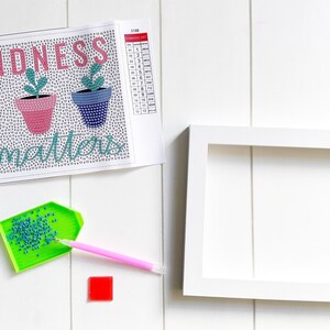 May include: A diamond painting kit with a white background and a colorful design of three potted plants with the text "Kindness matters". The kit includes a tray of diamonds, a pen, and a white frame.