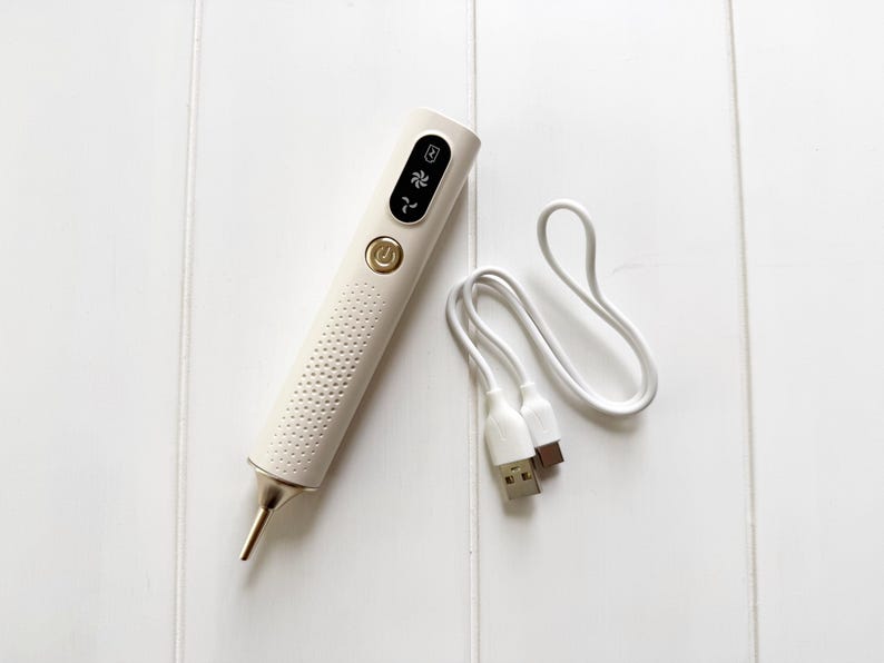 May include: A white beauty device with a gold-colored tip and accents, featuring a USB charging cable. The device has a textured body and a control panel with symbols. The background is a white wooden surface.