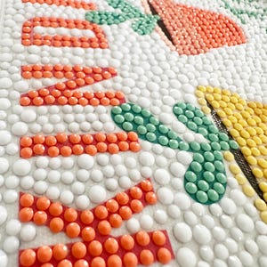 May include: Close-up of a colorful mosaic art piece with the word "Welcome" spelled out in orange beads. The background is white with small white beads.