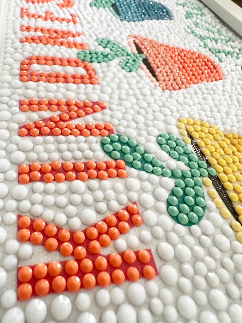 May include: Close-up of a colorful mosaic art piece with the word "Welcome" spelled out in orange beads. The background is white with small white beads.
