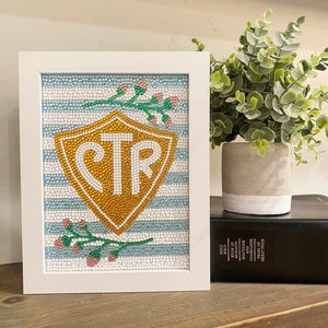 May include: A white framed picture with a blue and white striped background. A gold shield with the letters CTR is in the center of the picture. There are small green branches with pink flowers on either side of the shield.