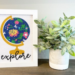 May include: A white framed beaded art piece with a colorful beaded globe and the word "explore" in black lettering.