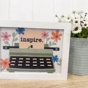 May include: A white framed picture with a green typewriter and the word "inspire" on a peach background. The typewriter is surrounded by colorful flowers.