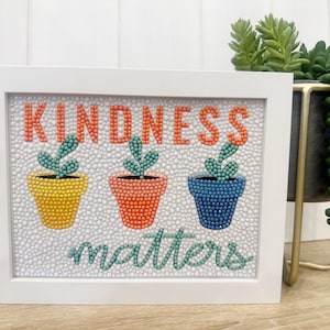 May include: White framed artwork with the words "Kindness matters" in green and orange. Three potted plants are depicted in yellow, orange, and blue.