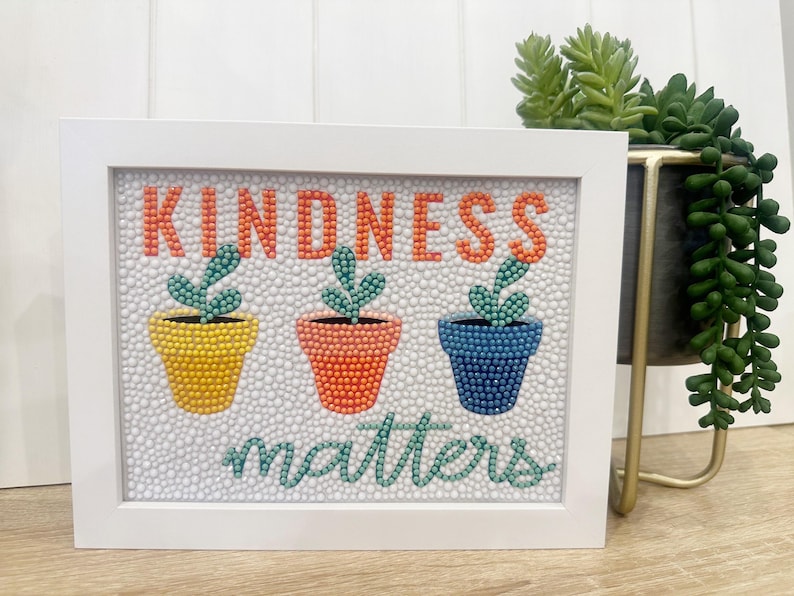 May include: White framed artwork with the words "Kindness matters" in green and orange. Three potted plants are depicted in yellow, orange, and blue.