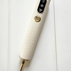 May include: A cream-colored beauty device with a gold-colored tip and accents. The device has a power button and a digital display with icons. The body of the device is perforated with small holes.