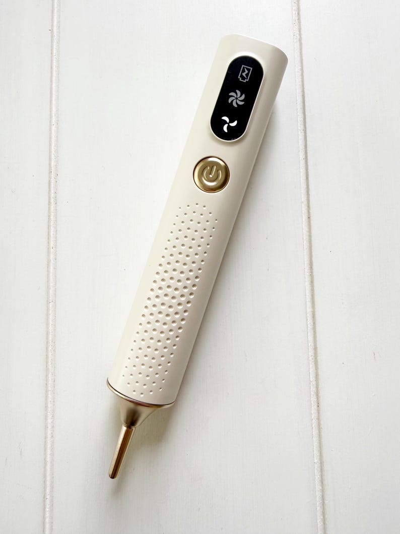 May include: A cream-colored beauty device with a gold-colored tip and accents. The device has a power button and a digital display with icons. The body of the device is perforated with small holes.
