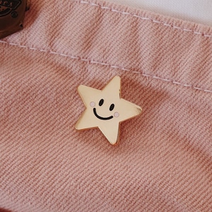 A pink denim overall with a gold star-shaped pin featuring a smiley face. The pin is attached to the front of the overalls.