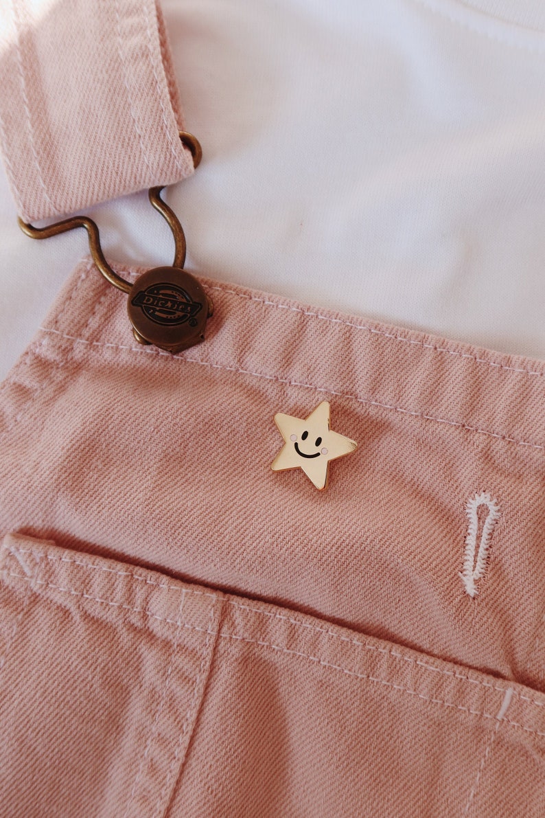 A pink denim overall with a gold star-shaped pin featuring a smiley face. The pin is attached to the front of the overalls.