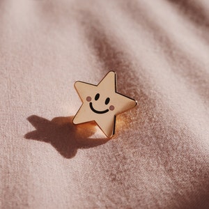 A gold star-shaped pin with a smiling face and rosy cheeks. The pin is on a light pink fabric background.