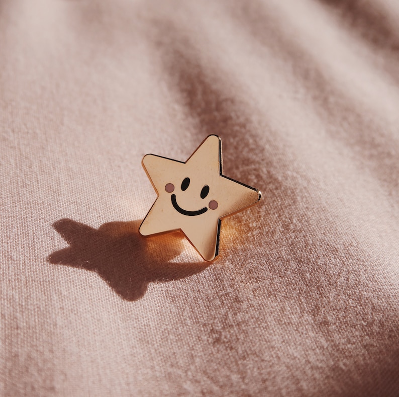 A gold star-shaped pin with a smiling face and rosy cheeks. The pin is on a light pink fabric background.