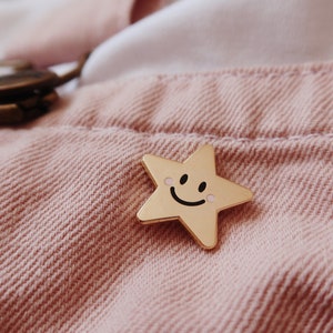A gold star-shaped pin with a smiling face and pink cheeks. The pin is on a pink fabric background.