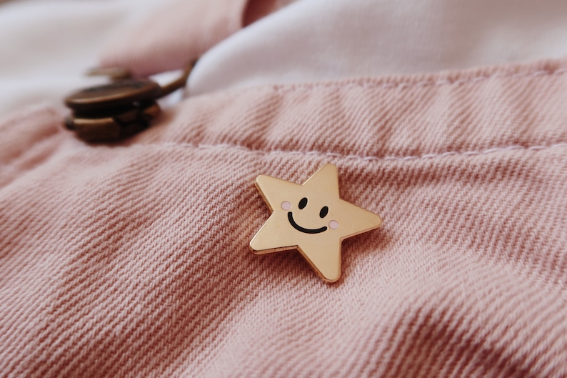 A gold star-shaped pin with a smiling face and pink cheeks. The pin is on a pink fabric background.