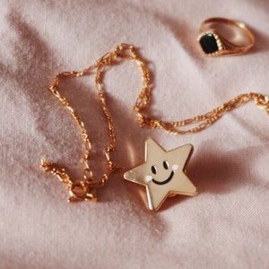 A gold chain necklace with a gold star charm featuring a smiley face. The necklace is lying on a pink fabric surface with a gold ring with a black stone.