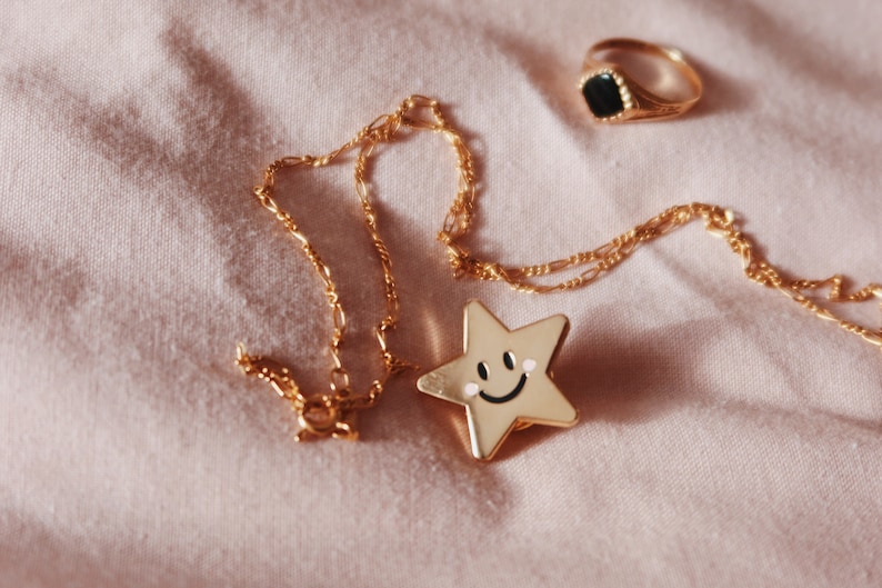A gold chain necklace with a gold star charm featuring a smiley face. The necklace is lying on a pink fabric surface with a gold ring with a black stone.