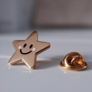 Gold-coloured star-shaped pin with a smiling face and pink cheeks. The pin has a gold-coloured back with a safety clasp.