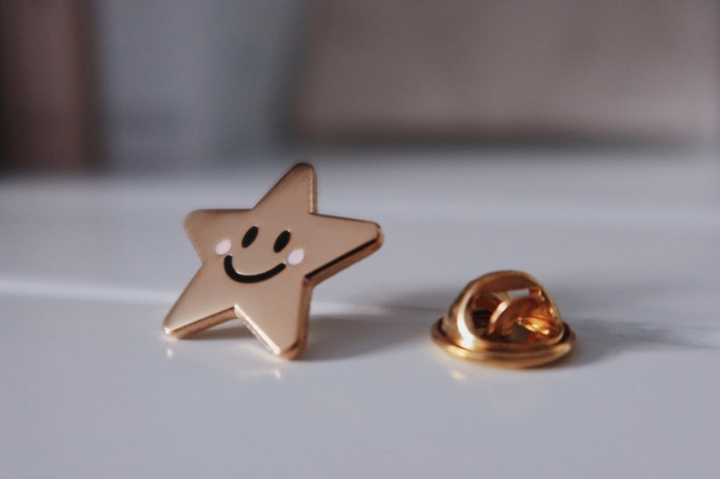 Gold-coloured star-shaped pin with a smiling face and pink cheeks. The pin has a gold-coloured back with a safety clasp.