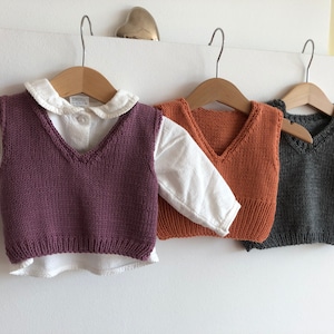 May include: Three knitted vests for children, one purple, one orange, and one grey. The vests are hanging on wooden hangers.